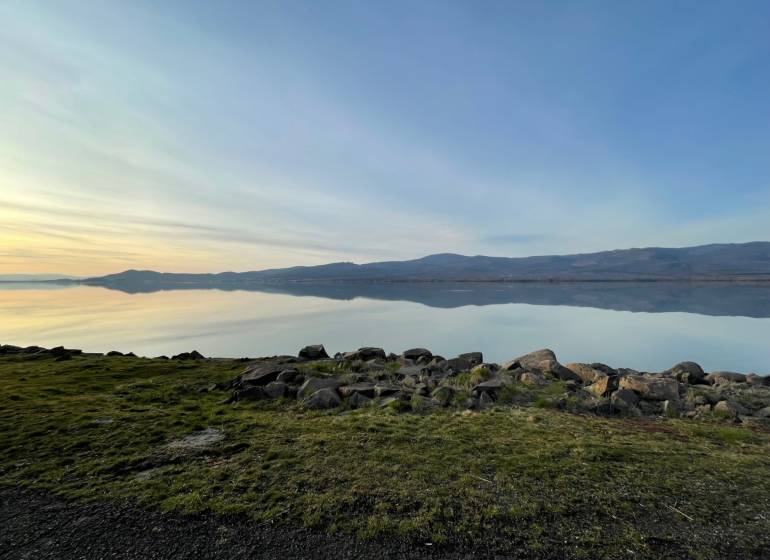 Nyugodt vízfelület, füves part és hegyek Vinné környékén. Lakóterületek.