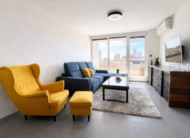 Nappali egy háromszobás lakásban sárga fotellel, kék kanapéval és falra szerelt TV-vel.