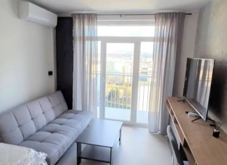 Garzonlakás kanapéval, televízióval és asztallal, fa dekorációjú padlóval, világos függönyökkel.