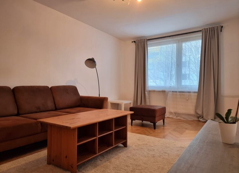 Nappali egy kétszobás lakásban, fa mintázatú padlóval, barna kanapéval és dohányzóasztallal.