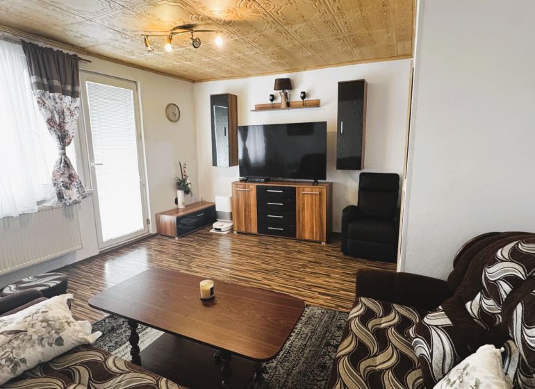 Nappali egy négy szobás lakásban, fa mintázatú padlóval és falra szerelt televízióval.