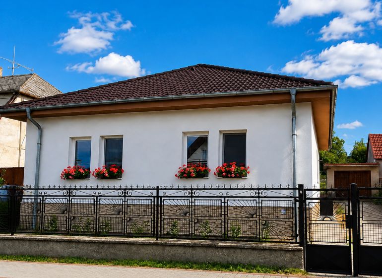 Családi ház Sellyén fehér homlokzattal, ablakvirágokkal és kontyolt tetővel.