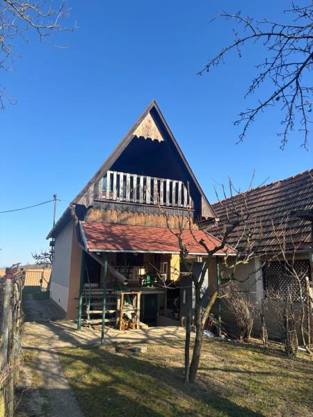 Ferde tetős, verandás házikó, amelyet kerti rekreációs területek vesznek körül.