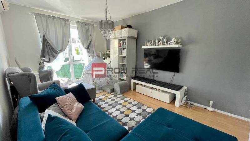 Nappali egy háromszobás lakásban kék kanapéval és falra szerelt televízióval.