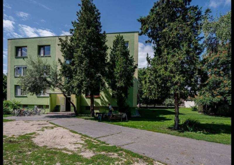 Egy fák által körülvett lakókonténer Csicsón, aszfaltút és kerékpárok mellett.