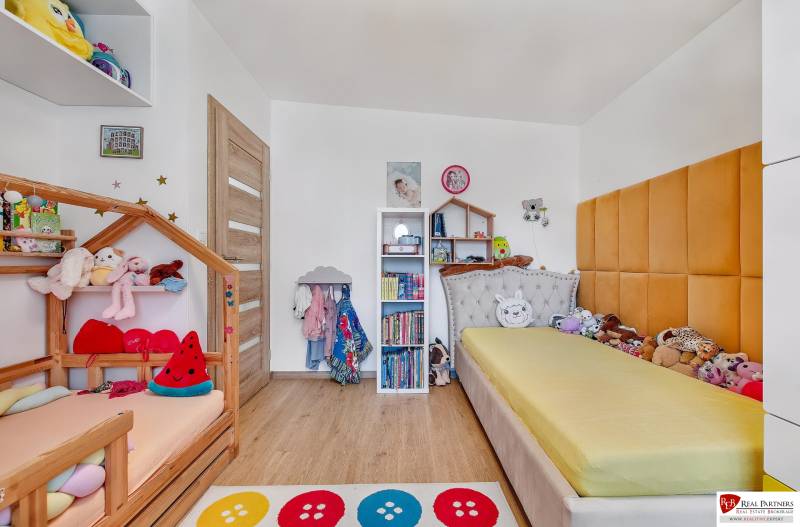 Gyerekszoba egy családi házban ágyakkal, polcokkal és játékokkal, világos fa padlóval.