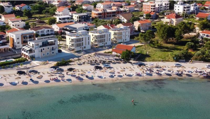 Strand napozóágyakkal és napernyőkkel, mögötte házak és épületek az üdülőhelyen.