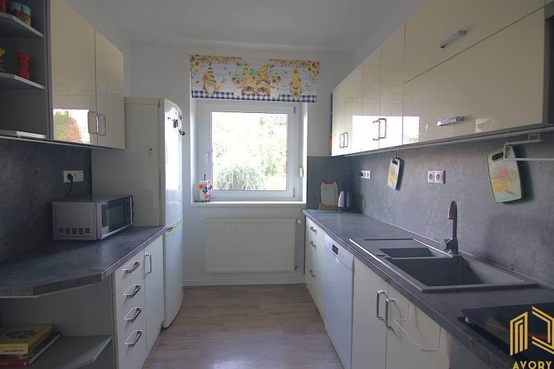 Családi ház konyhája fehér szekrényekkel, nagy ablakkal és háztartási gépekkel.