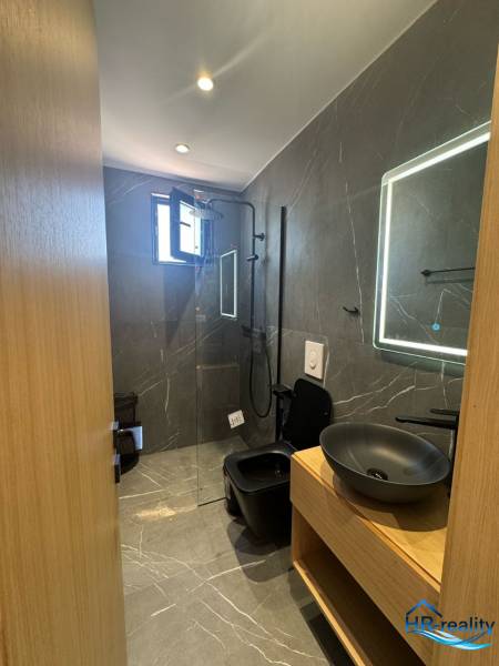 A nyaraló apartman fürdőszobája zuhanyzóval, fekete mosdóval és megvilágított tükörrel.