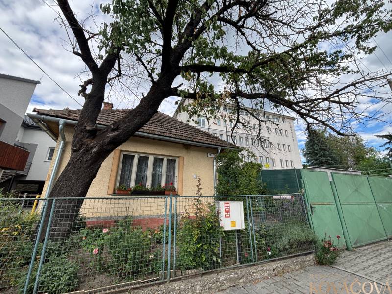 Családi ház Illaván kerített kerttel, fával és a háttérben szomszédos épületekkel.