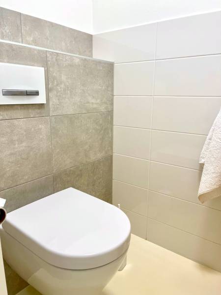 Egyszerű WC-k négyzet alakú öblítéssel egy kétszobás lakásban, fehér és szürke csempékkel.
