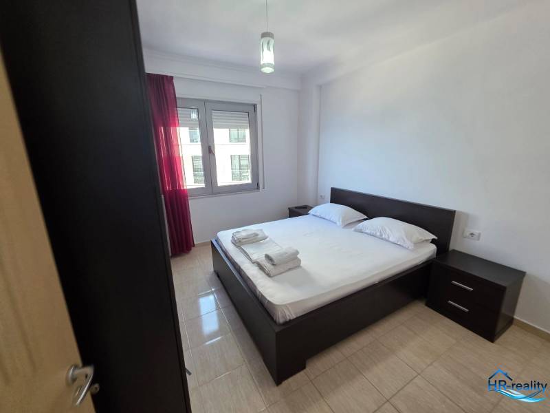 Hálószoba egy üdülő apartmanban, franciaággyal, éjjeliszekrényekkel és piros függönyös ablakkal.