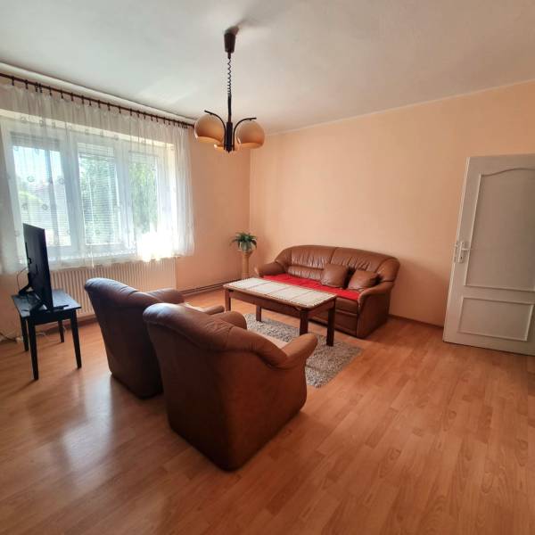 Családi ház nappalija kanapéval, fotelekkel, televízióval és szőnyegen álló faasztallal.