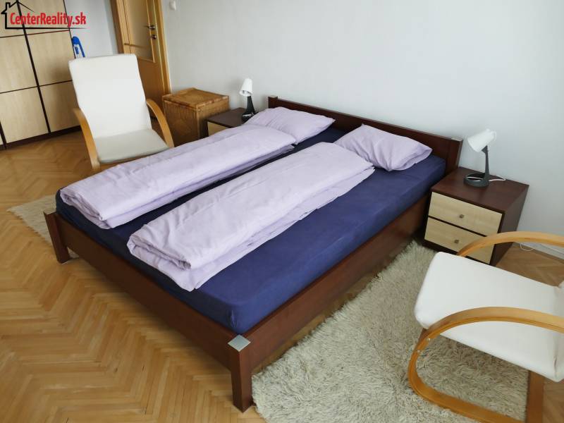 Hálószoba egy háromszobás lakásban, fa ággyal, székkel és fehér szőnyeggel.