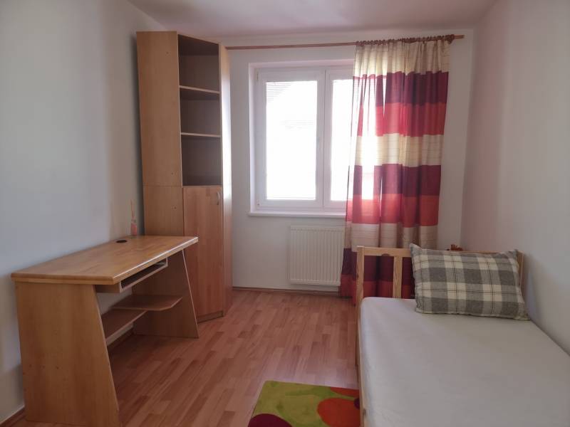 Egy szoba egy négy szobás lakásban, faasztallal, szekrénnyel és párnás ággyal.