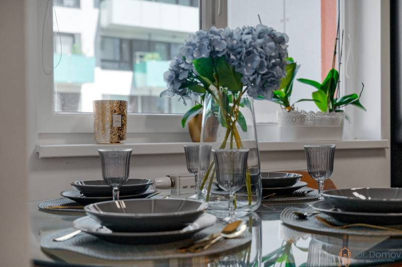 Elegánsan megterített asztal virágokkal egy kétszobás lakásban, üvegedények és evőeszközök.