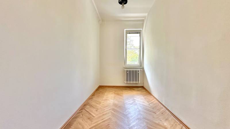 Szoba világos falakkal, fa padlóval, radiátorral és ablakkal egy háromszobás lakásban.
