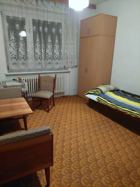 Egy családi ház szobája ággyal, szekrénnyel, asztallal és székekkel, meleg színekben hangolva.