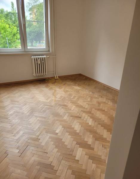 Szoba egy háromszobás lakásban, fa parkettával, nagy ablakkal, alatta radiátorral.