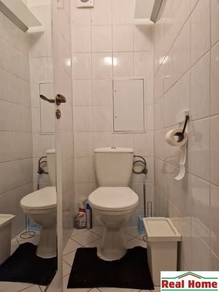 WC egy 2 szobás lakásban fehér csempékkel, fekete szőnyeggel és tisztítószerekkel.