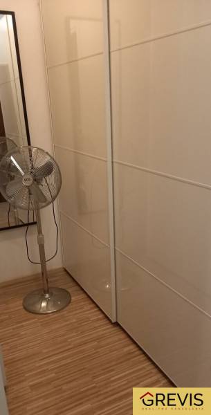 Gardrób egy kétszobás lakásban fehér tolóajtós szekrényekkel és álló ventilátorral.