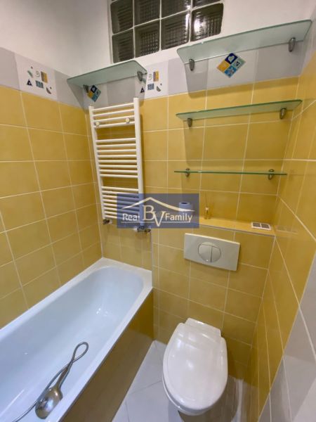 Fürdőszoba sárga csempékkel, káddal, WC-vel és üvegpolcokkal egy egyszobás lakásban.