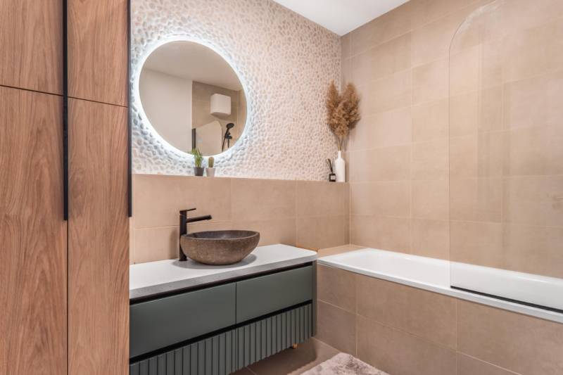 Fürdőszoba egy háromszobás lakásban mintás fallal, kerek tükörrel és káddal.