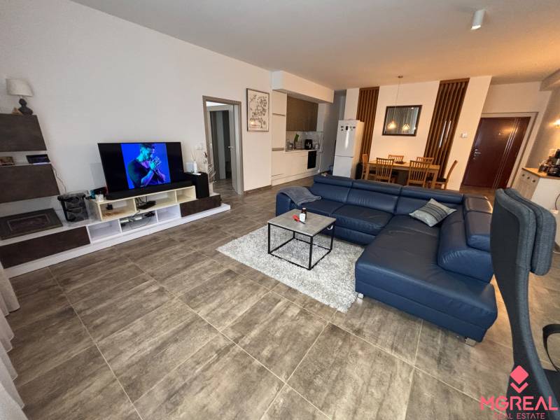 Nappali egy kétszobás lakásban sötét kanapéval és televízióval, fa mintás padlóval.