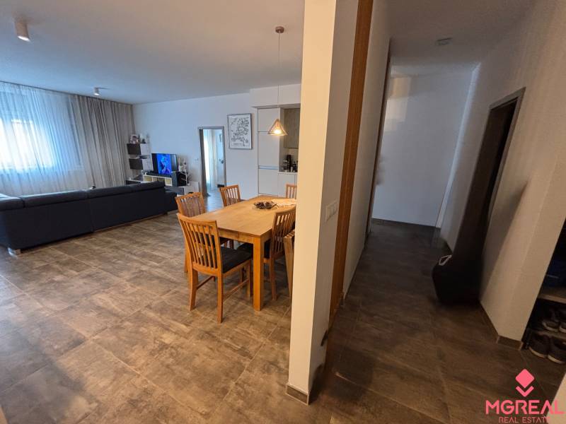 Étkezőasztal fa székekkel egy kétszobás lakásban. Része egy kanapé és egy televízió.