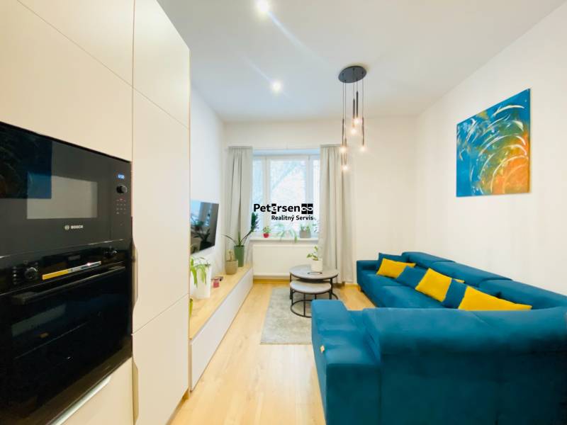 Nappali kék kanapéval, színes festménnyel és fa dekorációjú padlóval egy kétszobás lakásban.