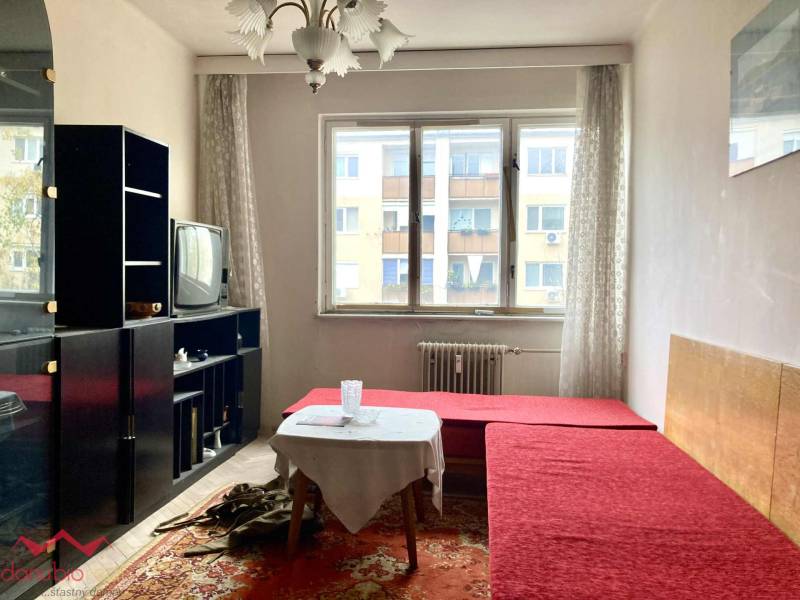 Nappali egy háromszobás lakásban piros kanapéval, szőnyeggel, régebbi televízióval, világos térrel.