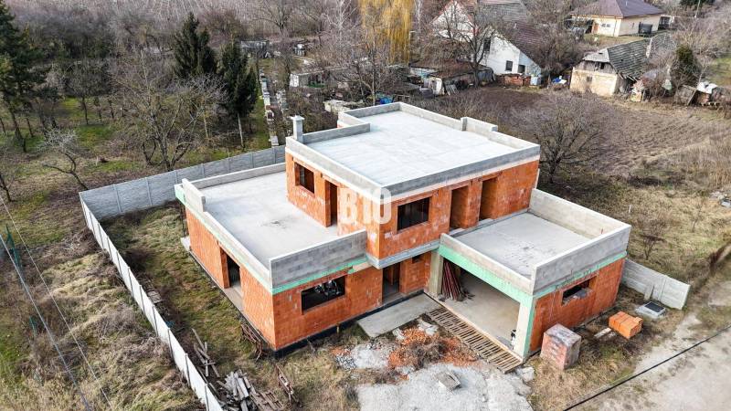 Építési projekt: családi ház, téglából készült szerkezet, tető nélkül, természeti környezetben.