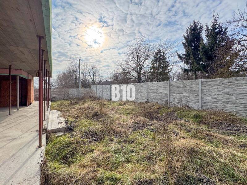 Családi ház építése füves telekkel és kerítéssel, felhős ég alatt.