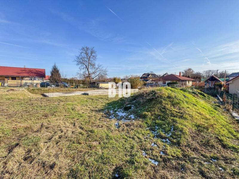 Építkezésre előkészített telek, havas fű, házakkal és tetőfedésekkel körülvéve.