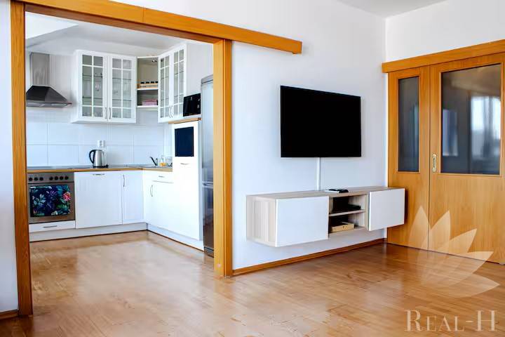 Nappali egy háromszobás lakásban, átjáróval a konyhába és falra szerelt televízióval.