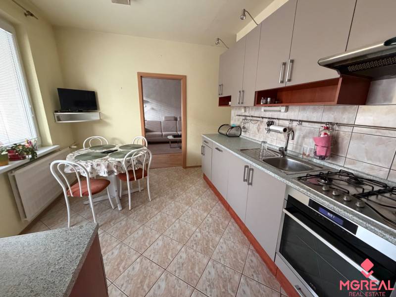 Konyha egy háromszobás lakásban sarok konyhabútorral, asztallal és falra szerelt televízióval.