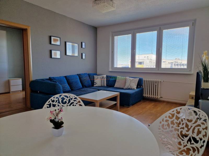 Nappali háromszobás lakásban kanapéval, fehér asztallal és fa dekorációjú padlóval.