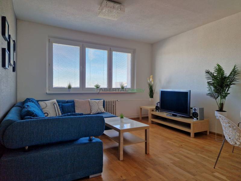 Nappali egy háromszobás lakásban kék kanapéval, televízióval és fa dekorációjú padlóval.