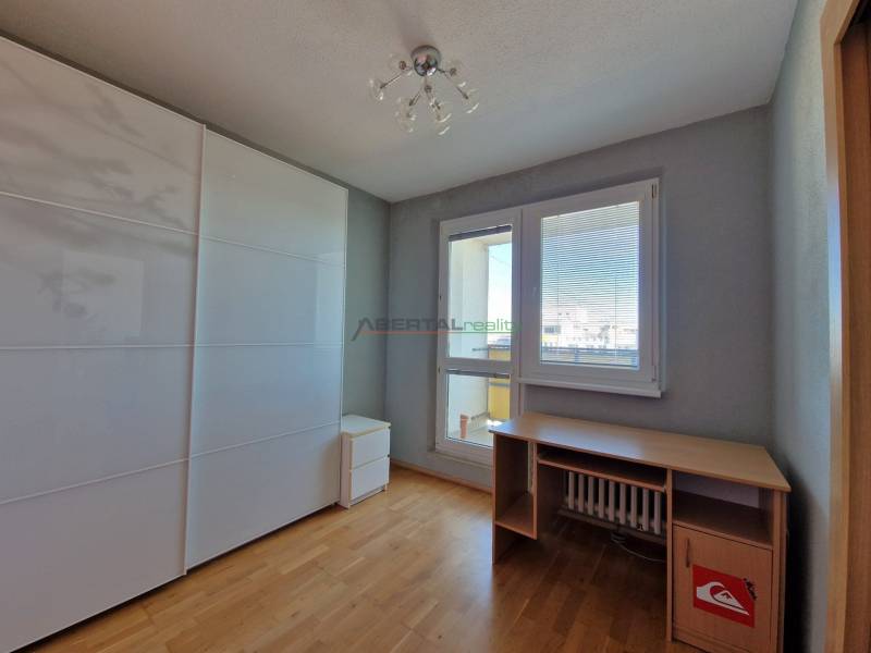 Szoba egy háromszobás lakásban, fa mintás padlóval, asztallal és szekrénnyel.