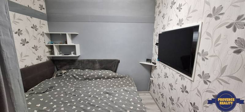 Hálószoba egy háromszobás lakásban, televízióval, virágos tapétával és polcokkal.