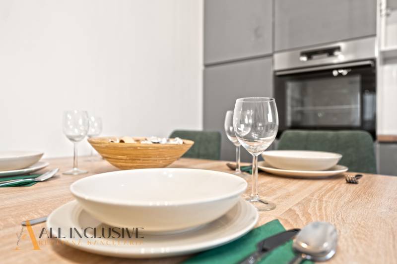 Étkezőasztal terítékkel és poharakkal, a háttérben egy családi ház konyhája.