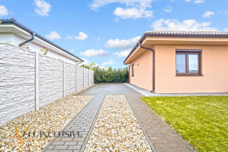 Családi ház Bősön, szilárd burkolatú bejárati úttal, kőkerítéssel és gondozott gyepfelülettel.