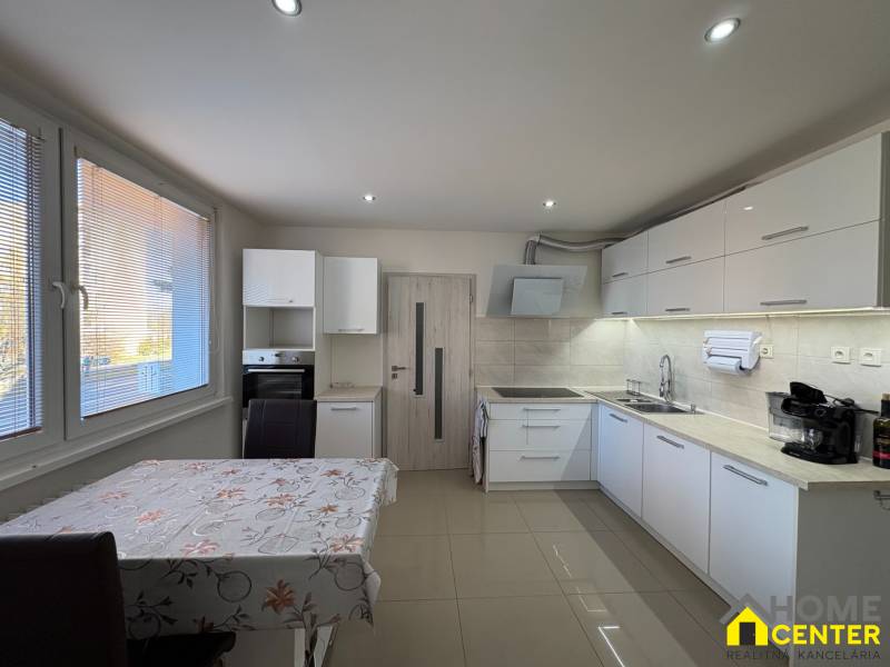 Konyha egy háromszobás lakásban fehér szekrényekkel és csempével, fa hatású padlóval.