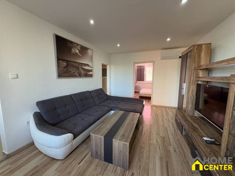 Nappali egy háromszobás lakásban sarokkanapéval és fa mintázatú padlóval.