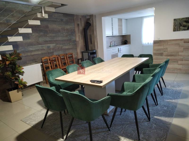 Ebédlő egy faházban nagy asztallal, modern székekkel és konyhasarokkal.