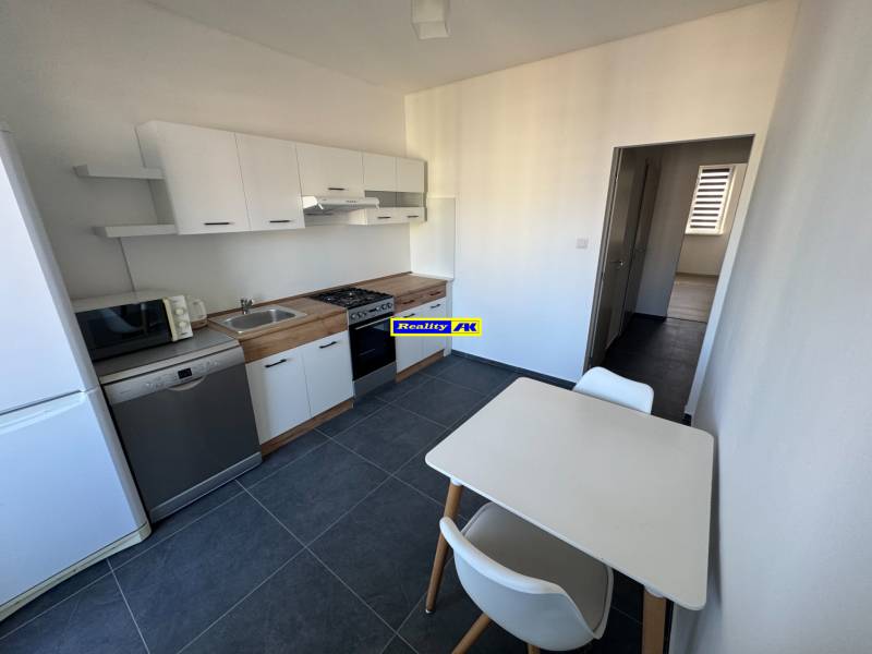 Konyha egy háromszobás lakásban fehér szekrényekkel és asztallal, sötét padlón.