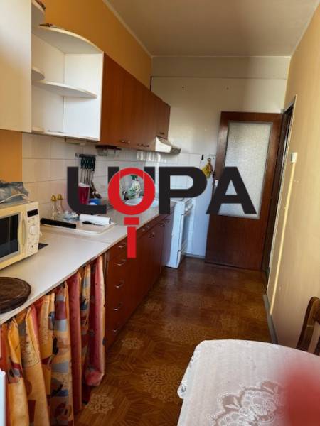 Konyha egy háromszobás lakásban, fa mintázatú padlóval, fehér szekrényekkel, étkezőasztallal.