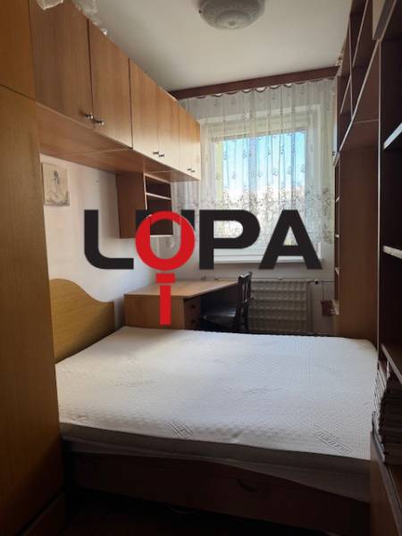 Hálószoba egy háromszobás lakásban, ággyal, szekrényekkel és csipkefüggönyös ablakkal.