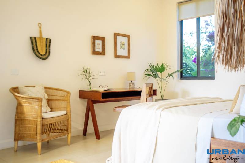 Világos hálószoba családi házban, rattan székkel és fa asztallal.