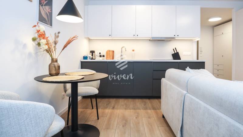 Konyha egy egyszobás lakásban fehér szekrényekkel, sötét munkalappal és fa hatású padlóval.